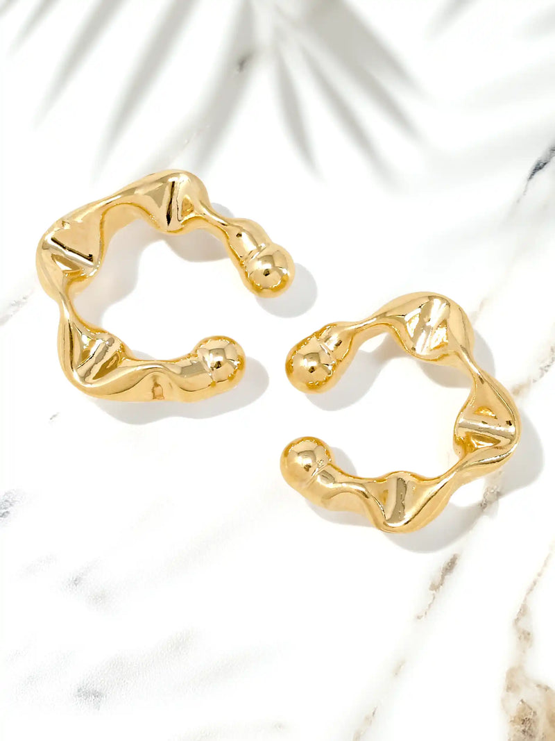 A pair of gold-toned abstract wavy hoop cuff earrings displayed on a white marble surface with soft palm leaf shadows.