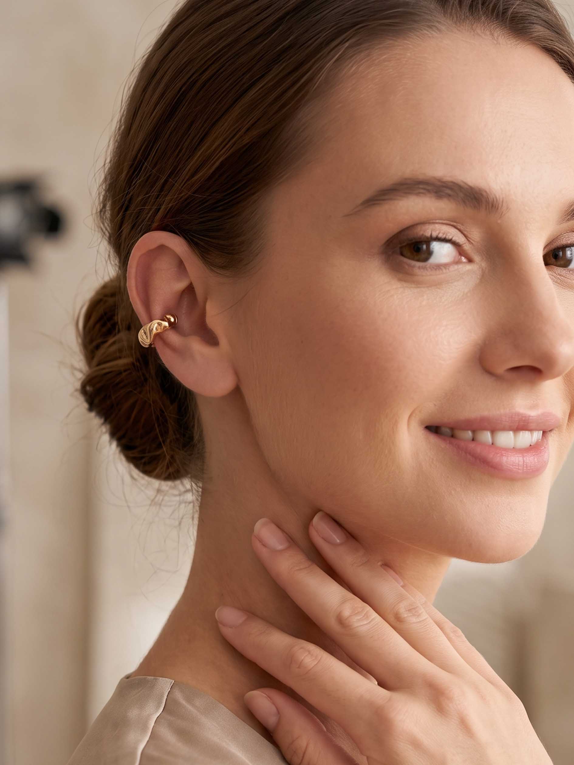 A close-up lifestyle shot of a woman with her hair in a low bun, wearing a delicate gold cuff hoop earring on her ear. She is touching her neck, with a softly blurred indoor background.