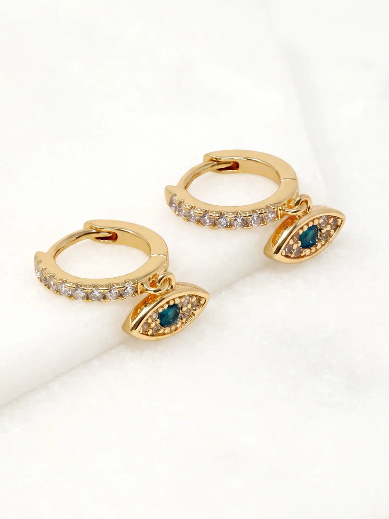 A pair of gold huggie hoop earrings with small clear pavé stones along the top band and dangling evil eye charms featuring dark blue-green gemstones in the center, displayed on a soft white textured surface.