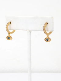 A pair of gold huggie hoop earrings with small clear stones and a dangling evil eye charm featuring a blue gemstone, displayed on a white jewelry stand.