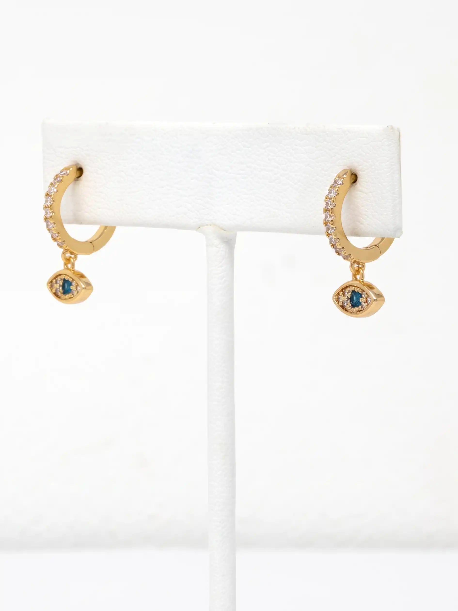 A pair of gold huggie hoop earrings with small clear stones and a dangling evil eye charm featuring a blue gemstone, displayed on a white jewelry stand.