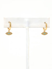 A pair of gold huggie hoop earrings with small evil eye charms featuring a turquoise blue center, displayed on a white jewelry stand against a white background.