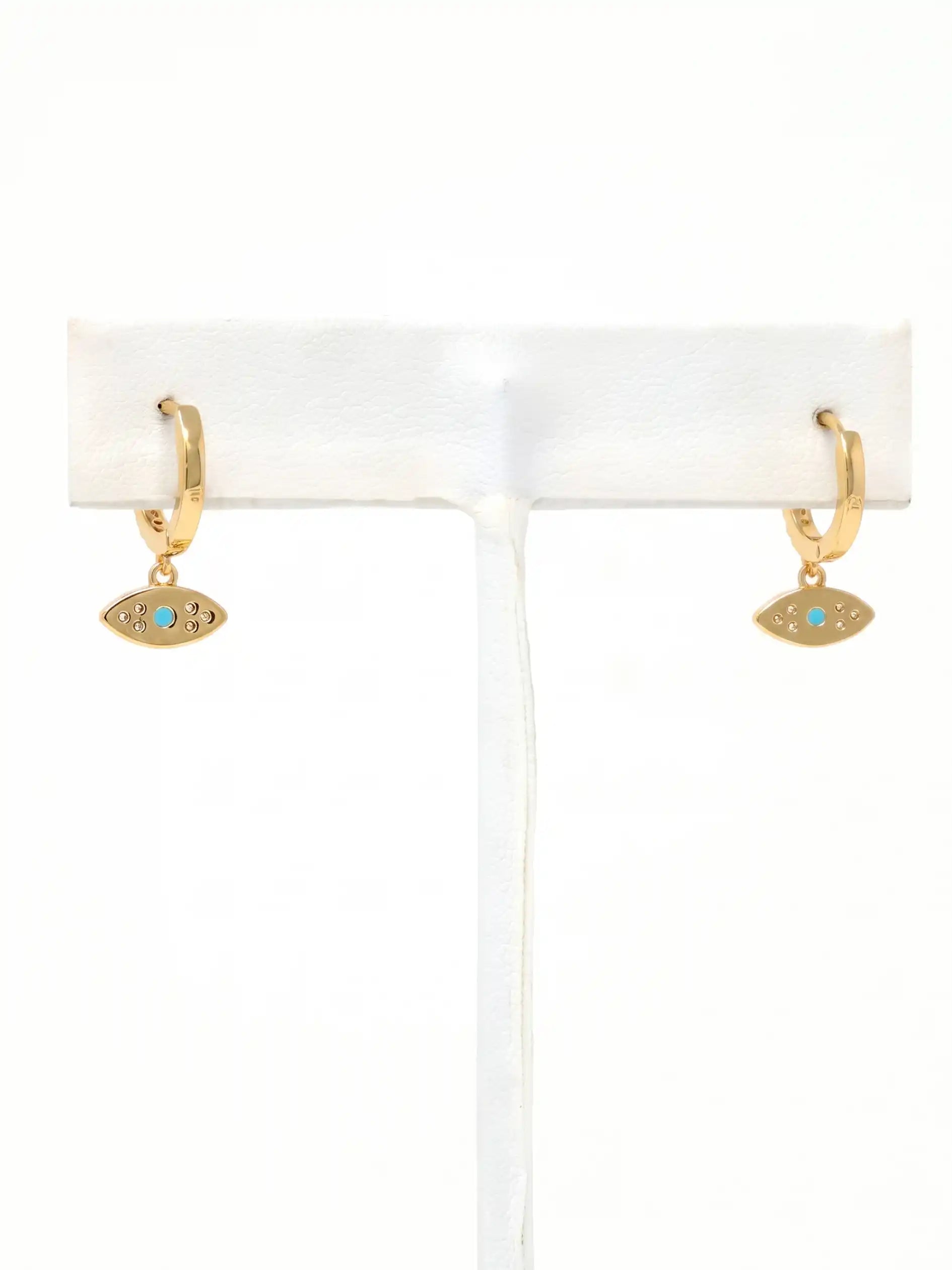 A pair of gold huggie hoop earrings with small evil eye charms featuring a turquoise blue center, displayed on a white jewelry stand against a white background.