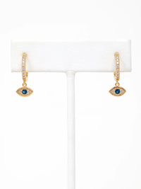 A pair of gold huggie hoop earrings with small clear stones and a dangling eye motif featuring a blue gemstone, displayed on a white jewelry stand against a white background.