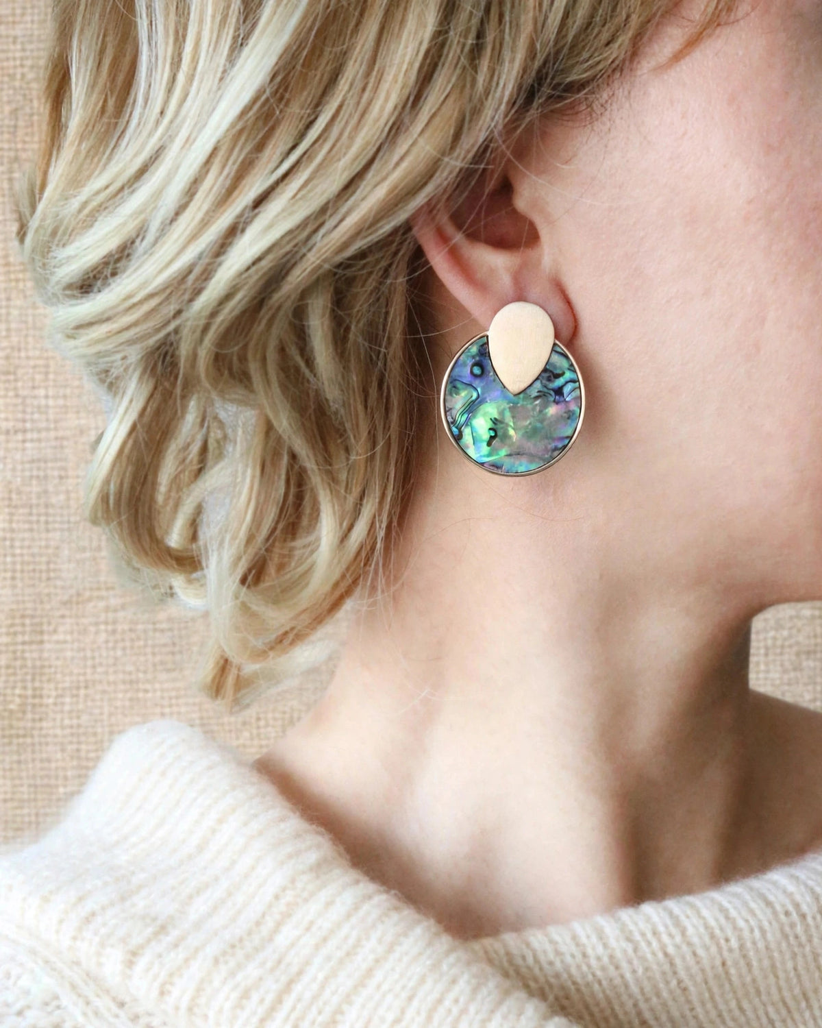 A close-up image of woman wearing a gold-tone round disc stud earrings with colorful iridescent abalone shell inlay in blue, green, and pink.