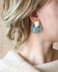 A close-up image of woman wearing a gold-tone round disc stud earrings with colorful iridescent abalone shell inlay in blue, green, and pink.