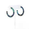 A pair of metal-mounted natural abalone shell hoop earrings. The iridescent abalone shells display beautiful shades of blue, green, black and purple, with a unique natural pattern visible from both sides. 