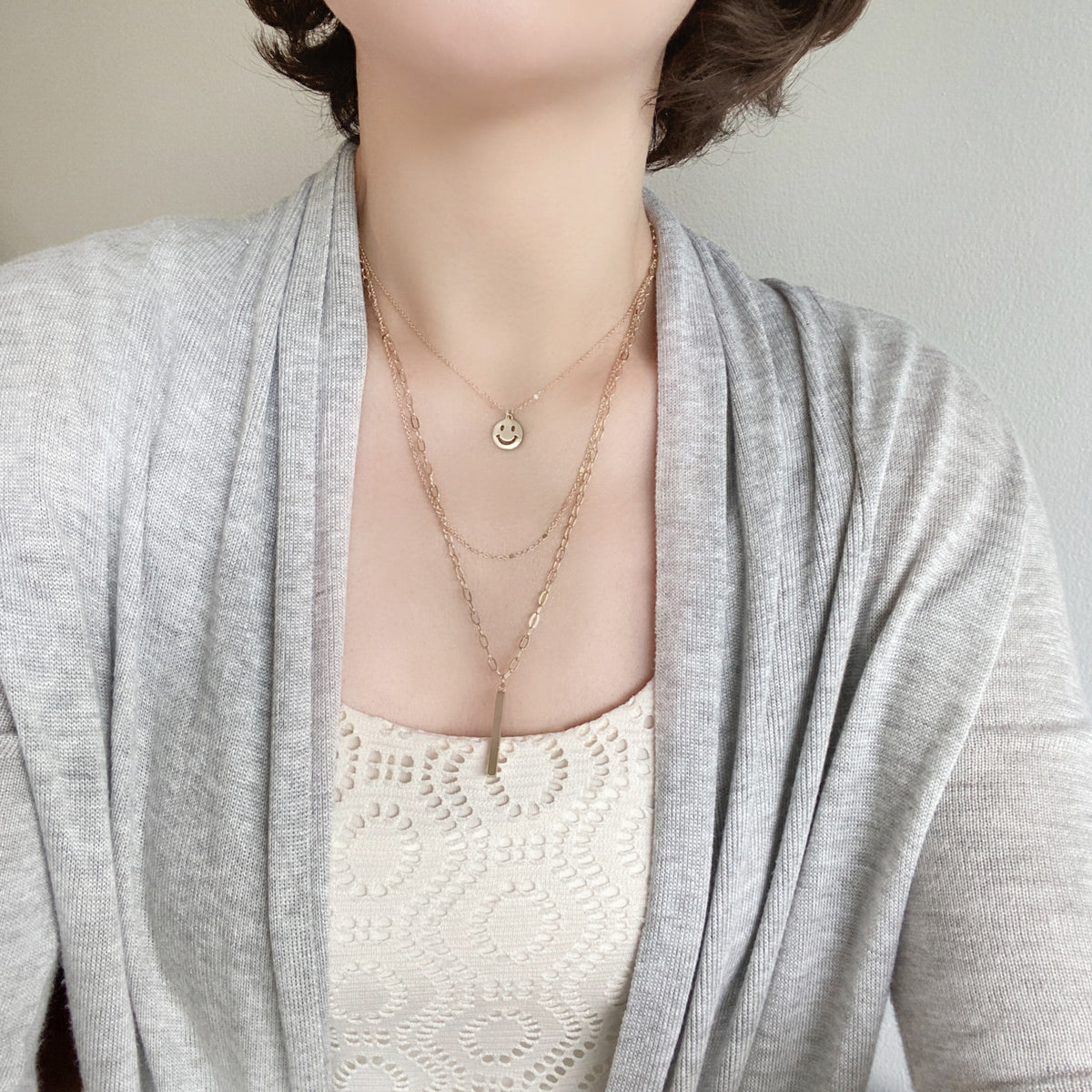 Wearing a set of triple layered necklace in gold with a small smiley face and a skinny bar charms.